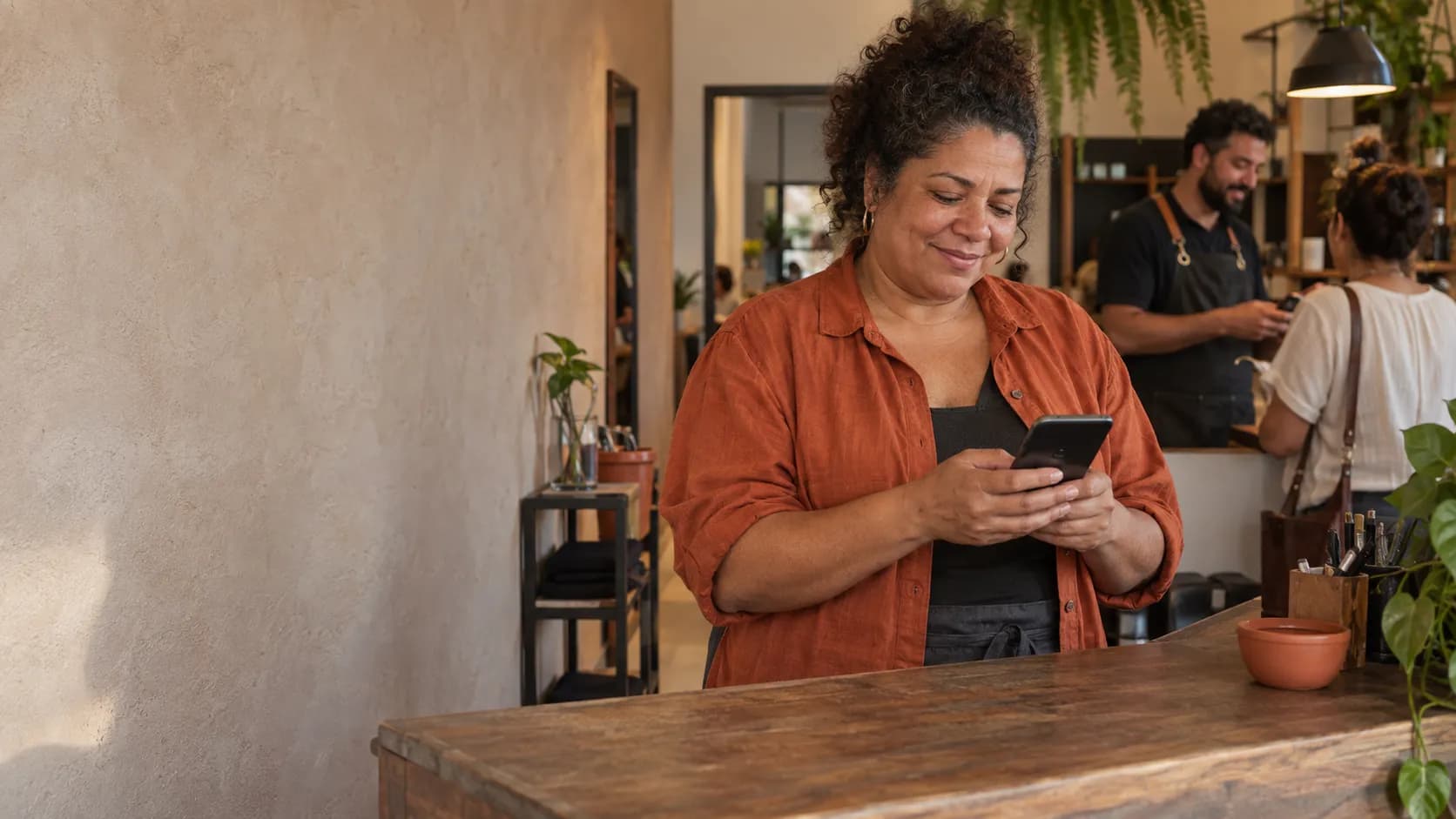 Dona de um pequeno negócio lê uma mensagem no celular atrás do balcão, enquanto um colega atende outra pessoa ao fundo.