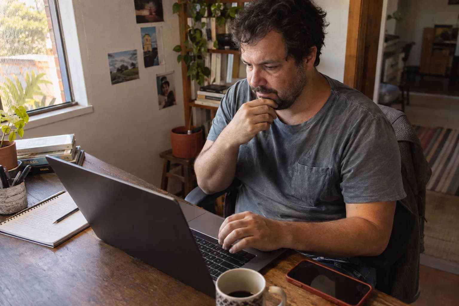 Em casa, um homem lê algo no notebook com a mão no queixo, com o celular e um café na mesa.